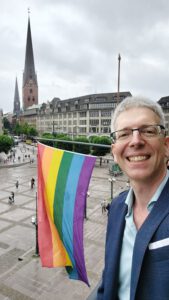 Foto vom Autor neben einer Regenbogenflagge, die am Hamburger Rathausmarkt gehisst ist.